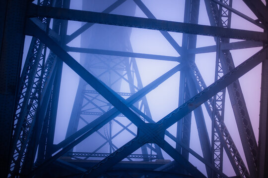 Steel structure of Dom Luis I Bridge in Porto disappearing into dense fog. Blue iron beams and geometric lines create a dramatic, abstract architectural composition.