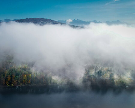 Aerial view of a tranquil lake shrouded in ethereal mist, with distant mountains peering through the clouds, Puidoux, Vaud, Switzerland.