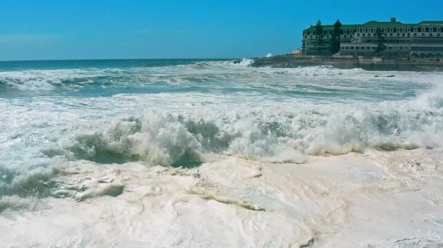 Aerial view over ocean with big rolling waves. Beautiful touristic city Ericeira on background of the ocean. Drone view of a Scenic slow motion ocean waves. Popular travel  place in Portugal, summer