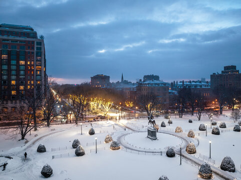 Aerial view of a snow-blanketed Boston Public Garden looking toward Commonwealth Ave, lights twinkling under a dark, twilight sky. Boston, Massachusetts, USA.
