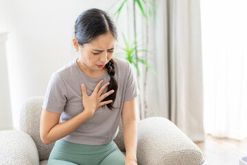 Obraz premium women holding chest in discomfort while sitting on sofa at home. Concept of chest pain, heartburn, anxiety, stress, breathing trouble, healthcare and consultation.