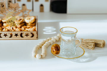 On the white table there is water in a glass, a date, a rosary, and a blurred tray with nuts.
