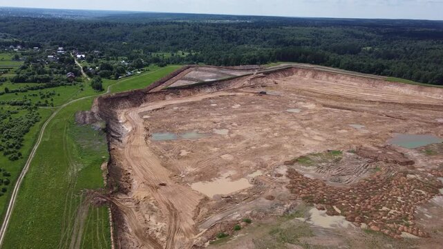 Drone soars over expansive sand pit expanding towards a distant village and lush green forests. Industrial quarry operations reshape the earth