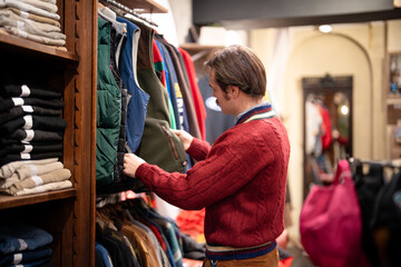 Fototapeta premium Man in old fashioned clothes choses different vests from display in vintage apparel store. Handsome guy looking at various clothing, mindfully picking items in pre owned goods store. Second hand.