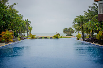 Obraz premium Infinity pool facing the beach on a rainy day, surrounded by tropical trees and lush greenery