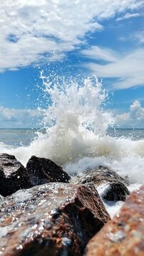Ocean wave crashes against dark wet rocks under blue sky