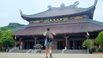 Young woman traveler with a backpack walking towards a large traditional buddhist temple building...