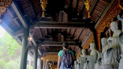 Naklejka premium Backpacker walking down a covered temple corridor lined with intricate stone arhat statues and ornate wooden carvings, evoking heritage, spirituality and quiet contemplation