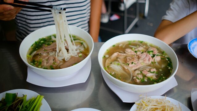 People enjoying authentic vietnamese pho soup with beef and rice noodles, using chopsticks to eat the traditional street food dish, served with fresh herbs and bean sprouts