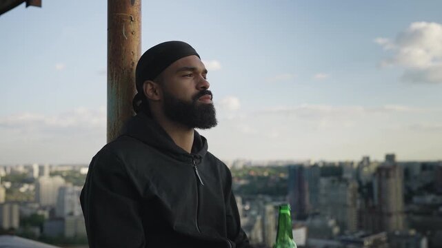 Young Black male drinking bottled beer on rooftop, thinking about life