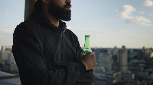 Pensive African American man drinking beer and looking at city from rooftop