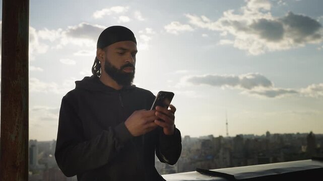 Young Black man in hoodie checking phone, setting up meeting on rooftop
