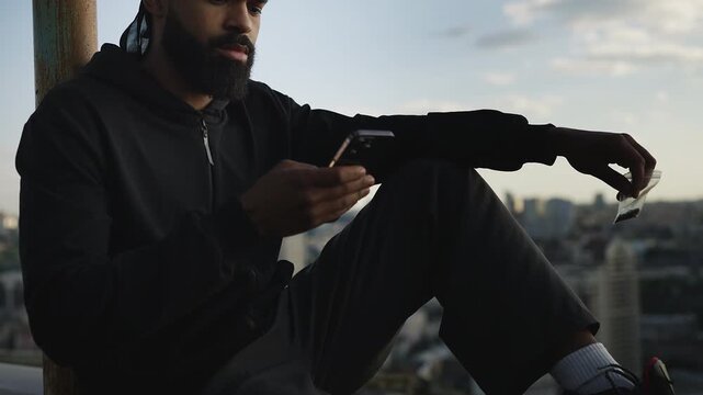African American man in hoodie selling marijuana online, texting on rooftop