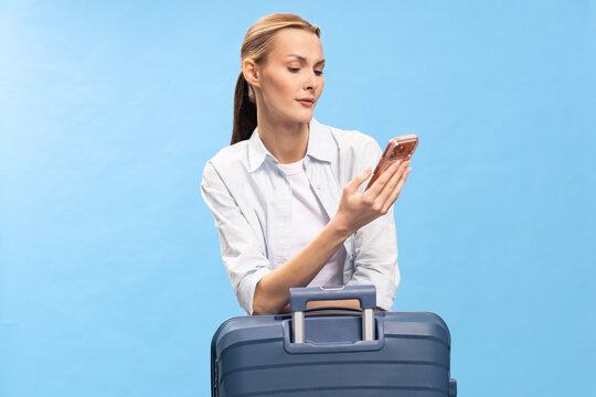 Young woman with suitcase looks at smartphone, preparing for travel. Casual outfit and blue background create concept of vacation, trip planning, and modern lifestyle.