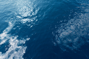 Aerial ocean background with deep blue water and subtle wave patterns, natural sunlight reflections creating abstract travel texture, clean modern composition