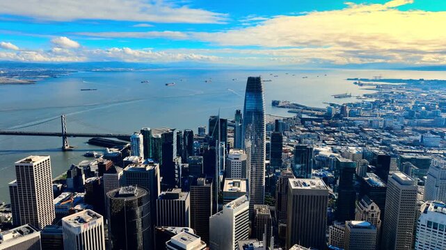 High angle aerial view looking towards the San Francisco Bay and Bay Bridge. The Salesforce Tower surrounded by mixed-use commercial buildings. Ships on the water under a partly cloudy sky.