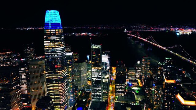 Aerial night view of San Francisco skyline featuring the illuminated Salesforce Tower and the Bay Bridge in the background. Sky is dark, and building lights are prominent.