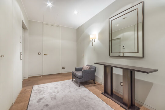 A gray chair is placed next to a console table in a simple hallway. A large mirror hangs on the wall above the table. Light comes from a wall lamp. The floor is wooden with a gray rug