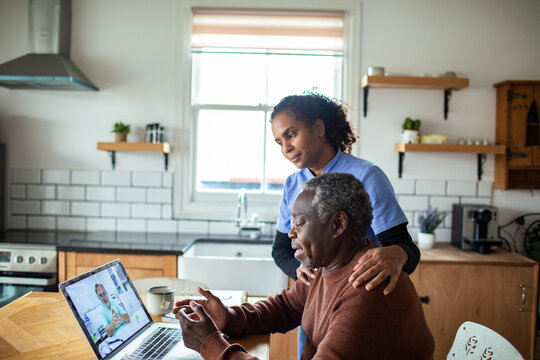 Caregiver supports senior during telehealth appointment in home kitchen