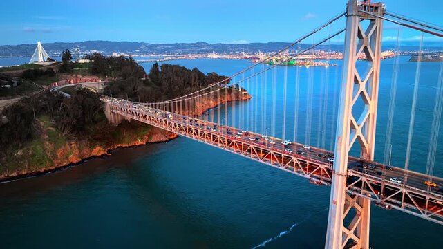 Aerial perspective looking down at traffic driving on the San Francisco Bay Bridge, approaching Yerba Buena Island. City skyline and port area visible in the background under a blue dusk sk