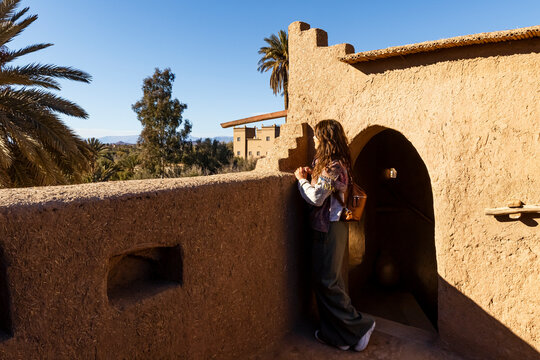 A woman looks out from a mud-brick building, with palm trees and a clear blue sky in the background. Kasbah Amridil,Marrakech,Morocco