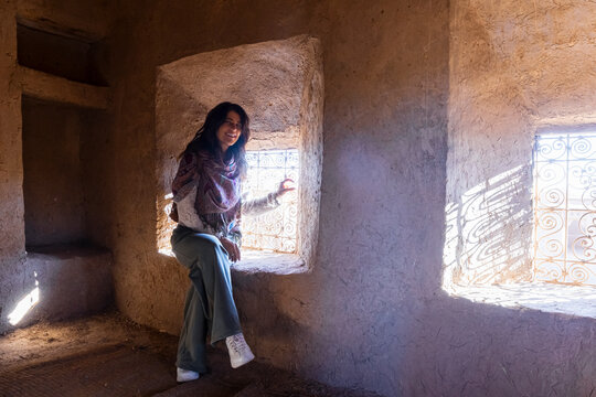 A smiling woman sits in a window of a mud-brick building, light streaming in. Kasbah Amridil,Marrakech,Morocco