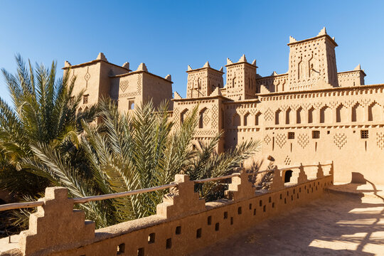 Exterior view of the Kasbah Amridil, a traditional earthen building with towers and geometric designs, framed by palm trees against a clear blue sky. Kasbah Amridil,Marrakech,Morocco