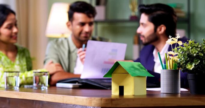 Indian couple finalizing home loan with real estate agent in office shaking hands while buying first dream house, cardboard home model in focus on table with blurred background