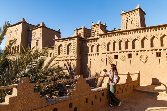 A woman stands on a wall overlooking a traditional mud-brick building with towers and geometric patterns under a clear blue sky. Kasbah Amridil,Marrakech,Morocco
