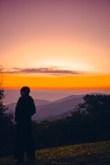 Fototapeta premium Blurred foreground silhouette of a woman standing on a mountain during sunrise. The sky is clear with warm tones of orange, yellow, and pink. Feeling fresh and free. Idea for travel with copy space.