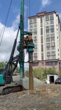  Green piling rig drills pile near high-rise building. Green excavator with drilling attachment on construction site, surrounded by dirt mound, small shed, car, and trees under partly cloudy sky. 
