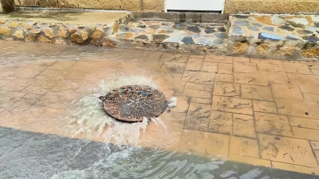 Water flows rapidly from a manhole cover on a tiled street, creating a splash as it spreads across the surface, with a stone border and building entrance visible in the background