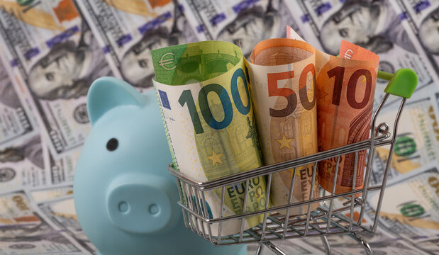 10, 50 and 100 euro notes in a shopping cart and a piggy bank against the backdrop of 100 US dollar notes