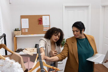 Fototapeta premium Diverse female colleagues examining garments on clothing rack in fashion studio with laptop and mug