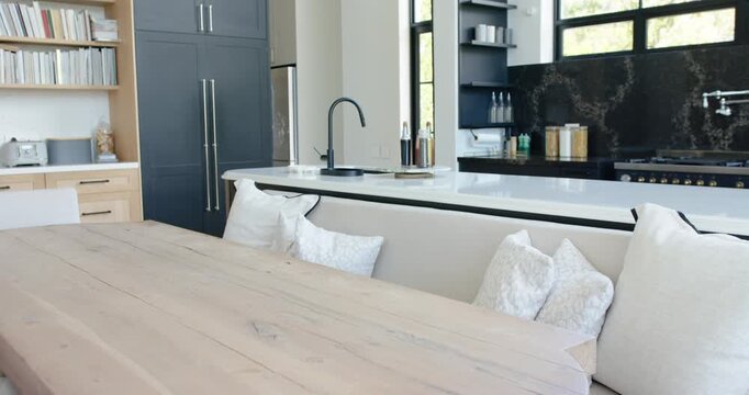 Camera is pulling back and panning right to reveal light wood table, bench, island, windows