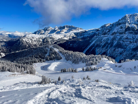 A vast alpine panorama photographed from the area of Giustino shows a winter landscape of the Dolomites under bright, clear sunlight.