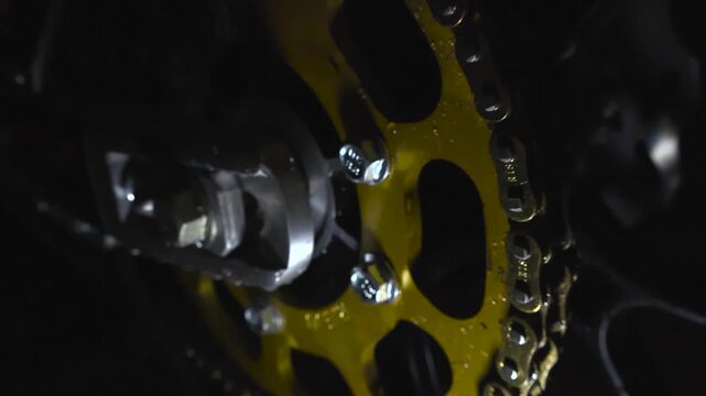Moody close up of gold anodized motorcycle chainring with black chain and rain droplets under dramatic low light conditions.
