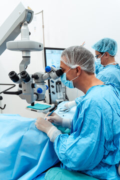 A specialized ophthalmic surgeon uses a high-precision microscope during a delicate surgical procedure in a sterile operating room with medical staff.