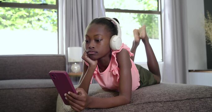 African American girl child lying on chaise switching from headset listening to tapping pink phone