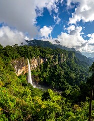 Tall waterfall cascades from a cliff face, surrounded by green forest and mountains under a cloudy sky