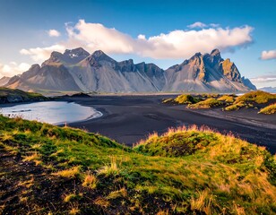 Dramatic peaks backdrop to black sand, inlet, and green tundra under a blue sky and clouds
