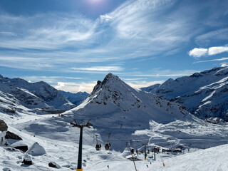 Fototapeta premium Whispers of Winter in the High Alps, A vast snowy slope stretches toward rugged alpine peaks, partially veiled by drifting clouds under a deep blue sky. The scene captures the serene beauty and raw ma