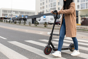 A young woman in a beige coat walks with an electric scooter through a pedestrian crossing in the city. Eco-friendly transport.