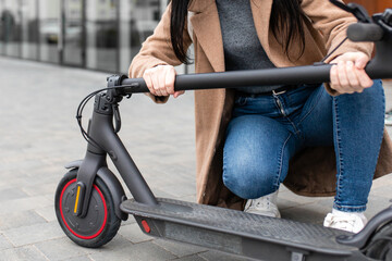 Obraz premium Young woman in a beige coat folds an electric scooter in the middle of a city street. A person uses an environmentally friendly form of transport in the form of an electric scooter.