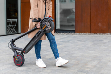 Young woman in a beige coat carries an electric scooter in his hands on the street. A person uses an environmentally friendly form of transport.
