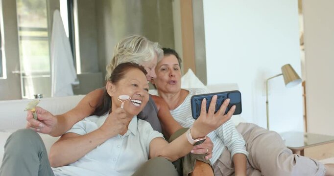 Diverse women friends holding phone, posing for selfie in living room, using roller and smiling