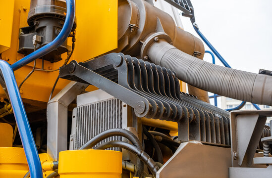 Close-up of industrial hydraulic oil cooler and protective steel grille on yellow heavy machinery. Detailed view of hydraulic hoses, radiator, and engine air intake system on a construction vehicle.