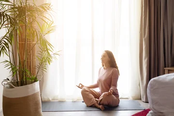 Fotobehang Lotusbloem Adult caucasian woman meditating in lotus position on yoga mat by window, female practicing meditation, calm hotel room wellness lifestyle concept, copy space  © Anastasiya