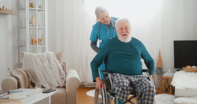 Nurse pushing retired senior man with disability in wheelchair at home, smiling and communicating warmly. Concept of mobility support, home care and compassionate healthcare.