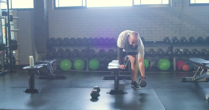 African American man at gym in shorts reaching for dumbbell bracing on bench pulling rows training